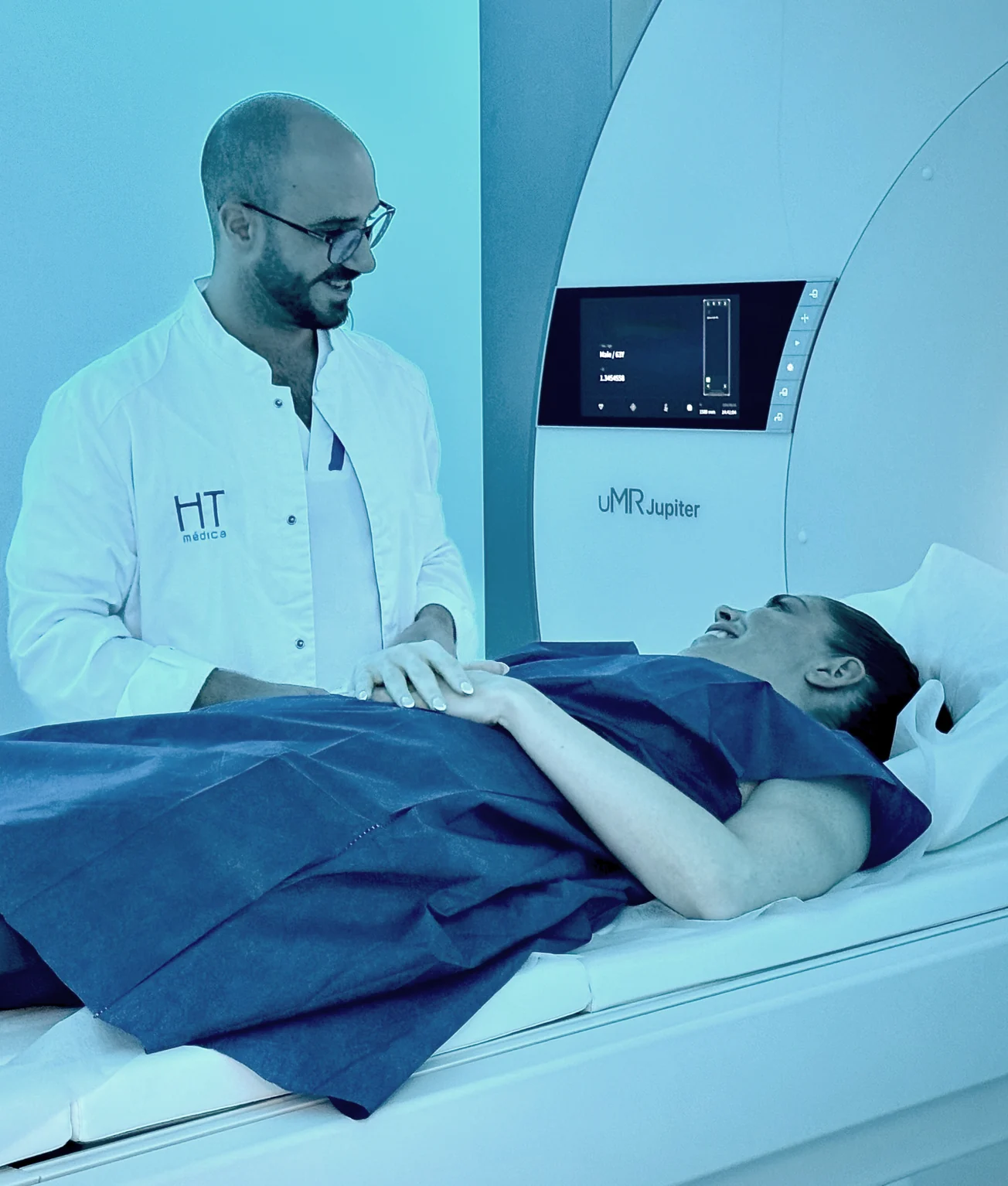 Paciente tumbada y sonriente en la mesa de la resonancia RM 5T de HT Médica, con una enfermera al fondo de la sala.